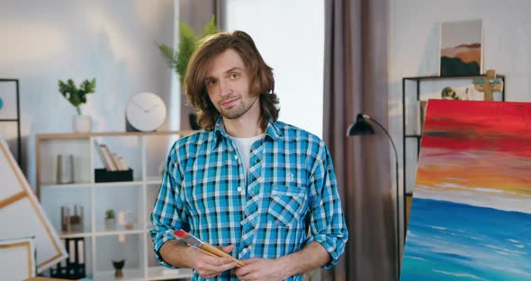 Male artist Holding Paintbrushes in Hands Standing Near Abstract Beautiful Picture Drawn on canvas alt