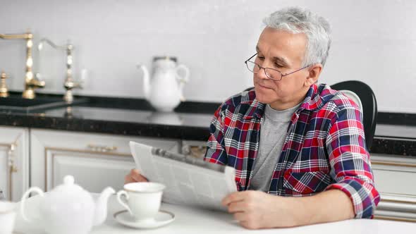 Aged Domestic Man in Glasses Reading Newspaper During Breakfast at Kitchen alt