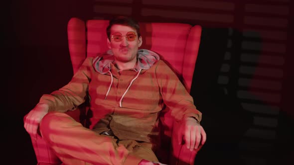 Young man in glasses sitting in armchair in dark room with falling red light and shadow blinds. alt
