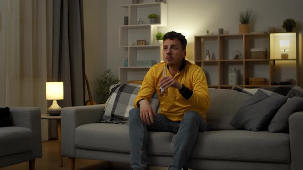 Young Caucasian Man Sitting on the Couch in the Living Room While Watching Sport Game on TV and alt