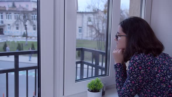 A Brunette Girl in Glasses Stands at the Window in Anticipation Side View Copy Space alt