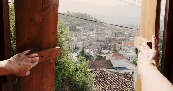 Girl Opening Wooden Sash Windows Overlooking to Old Town Berat in Morning Summer Sunlight alt