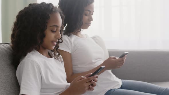 Little African American Girl and Her Young Mother Networking in Smartphones Sitting on Sofa and alt