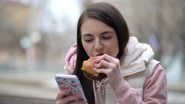 Happy Woman Eating a Burger and Using Smart Phone on the Street