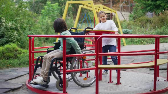 Wide Shot Positive Pretty Girl Spinning Merry Go Round with Disabled Cute Boy in Wheelchair alt