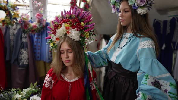 Charming Young Woman in Red Dress with White Embroidery Sitting Indoors As Friend Adjusting Head alt