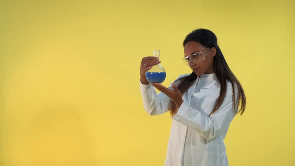 Black Female Scientist in Lab Coat Looking on Flask with Experimental Liquid on Yellow Background. alt