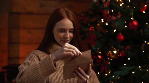 Close-up View of Smiling Young Woman Writing Christmas Card with Wishes on Background of Xmas Tree alt