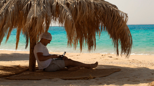 счастливый человек с зонтиком сидит на пляже с ноутбуком