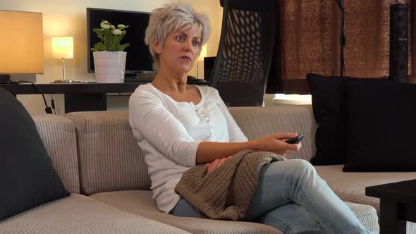 A Middle-aged Woman Sits, Watches TV and Switches Channels with a Remote Control alt