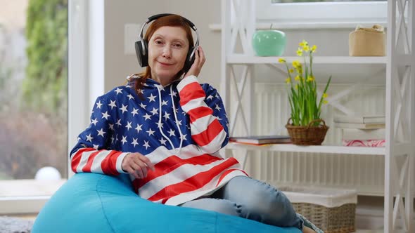 Happy Mature Woman in Hoodie Colored As USA Flag Looking at Camera Smiling Sitting on Bag Chair alt