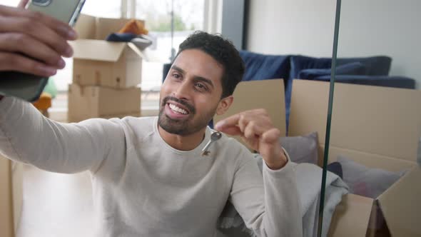 Man Holding Keys With Mobile Phone Moving Into New Home Making Video Call Surrounded By Boxes