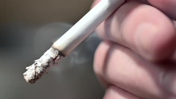 A man's hand holds a smoldering cigarette. Blue smoke comes from a cigarette.