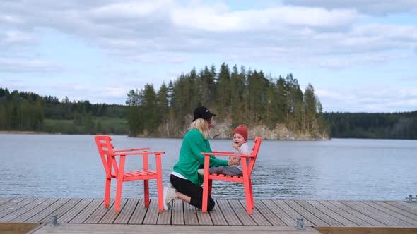 mom and little son are sitting on red chairs on the pier by the lake