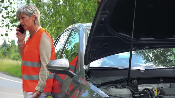Middle Aged Woman Wears Warning Vest and Phones with Smartphone Next To the Broken Car alt