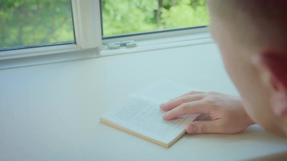 Young Man Reading a Book Sitting at an Open Window on a Sunny Day Back View alt