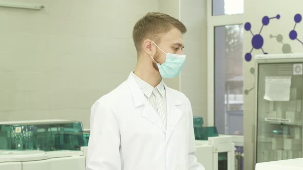 A Serious Lab Assistant Stands in the Middle of the Laboratory and Turns His Head Into the Camera alt