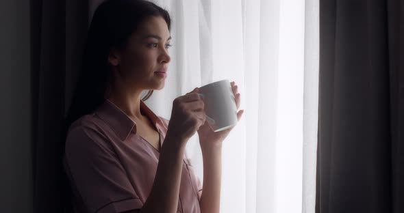 Woman Stands Near the Window with the Curtains Open Drinking Tea From a Mug
