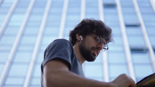 Delivery Man with Yellow Backpack Talk to a Client on the Street Using Earphones Low Angle View alt