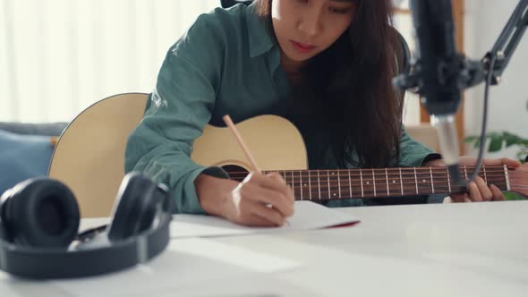 Happy asia woman songwriter play acoustic guitar listen song from smartphone think and write notes.