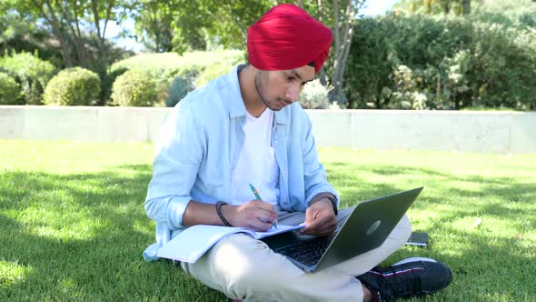 Serene Indian Male Student in Traditional Turban Using Laptop for Online Studying Sitting Outdoors