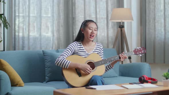 Asian Woman Composer With Paper And Notebook On Table Singing And Playing Guitar At Home alt