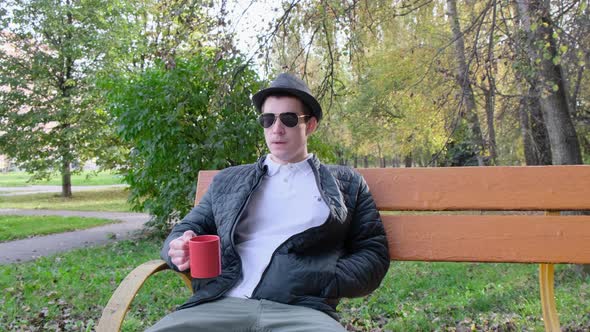 a young man in sunglasses is drinking tea in a glass on a bench. 4k