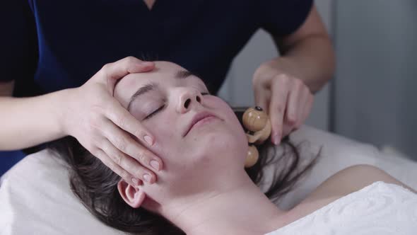 Woman Massagist Using a Wooden Roller Under the Jawline of Her Female Client alt