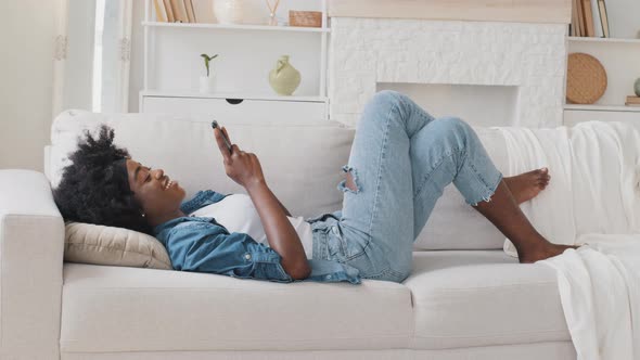 Lazy Smiling African Young Woman Surfing Social Media Shopping in App Playing Mobile Game Texting alt