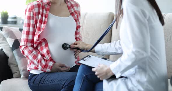 Doctor Listens with Stethoscope to Belly of Pregnant Woman at Home alt