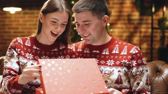 Romantic Couple Opening a Present Gift Box in the Evening Near Decorated Xmas Tree alt