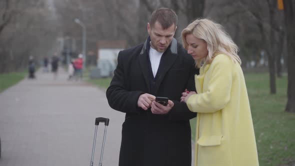 Adult Caucasian Couple Standing in Park with Suitcase and Using Smartphone alt