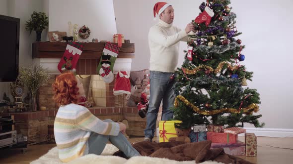 Wide Shot Portrait of Relaxed Caucasian Man in Party Hat Decorating Christmas Tree and Stepping on alt