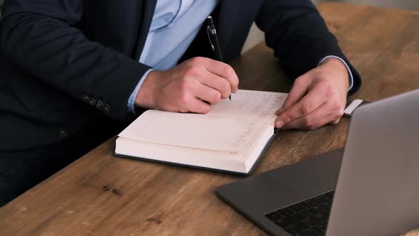 Side View of Businessman in Suit Filling Out Planner or Notepad Next to an Open Laptop Against of