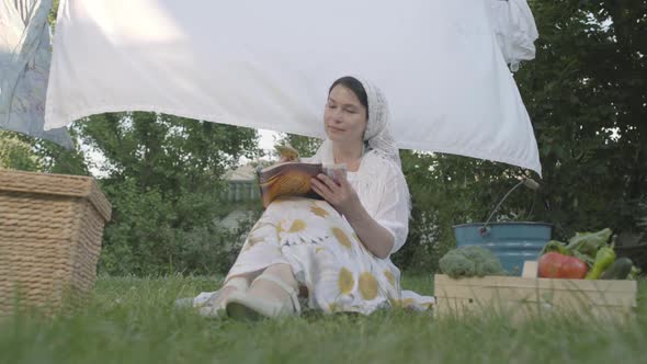 Adorable Woman with a White Shawl on Her Head Resting in the Garden Sitting on the Grass alt