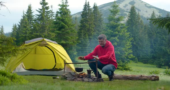 Bearded Male Tourist Warms His Hands Over a Bonfire in the Mountains alt