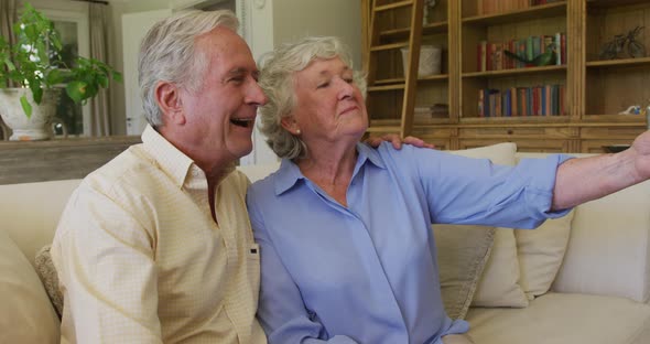 Smiling caucasian senior couple taking a selfie from smartphone sitting on the couch at home alt