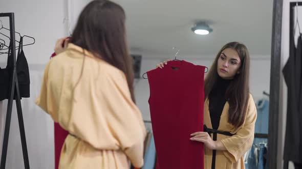Reflection in Shop Mirror of Focused Caucasian Beautiful Woman Trying on Red Sleeveless Shirt alt