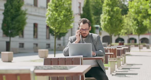 Young Man Wearing Glasses with Mustaches and a Beard is Sitting at the Table in the Square is alt