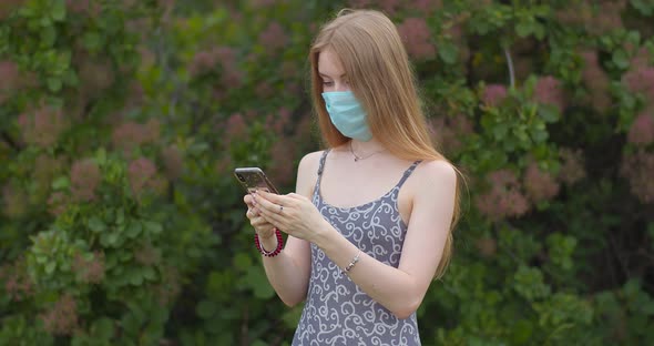 A young girl in a protective medical mask looks at the phone and sends messages