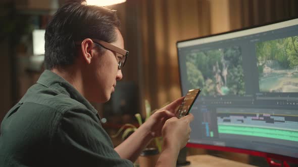 Asian Male Video Editor Works And Using Mobile Phone With His Personal Computer alt