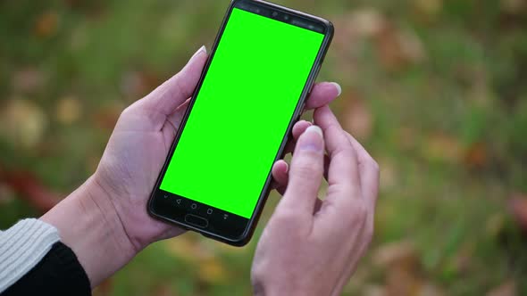 Woman's Hand holding mobile phone with green screen on autumn blurred background