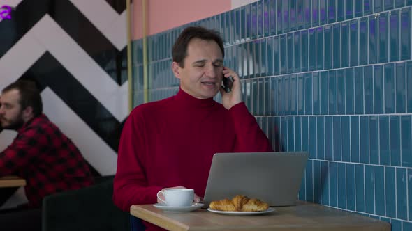 Smiling Middleaged Man Talking on Mobile Phone Sitting with Laptop in Cafe