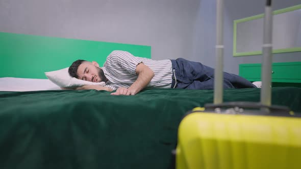Wide Shot of Young Handsome Man Sleeping on Comfortable Cozy Bed with Packed Yellow Travel Bag at alt