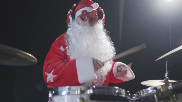 Cheerful Musician Dressed As Santa Claus Plays Drums on a Black Studio Backlit Background alt