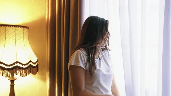 Young happy smiling woman looks out from a window of a hotel room. Large windows  alt