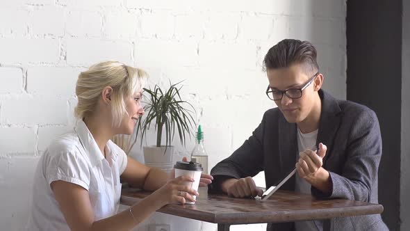 Man and Woman Sitting at the Table in Cafe. They Talking and Watching on Tablet alt