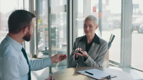 Smiling Satisfied Middle Aged Woman Sitting at the Table with Car Seller Shaking alt