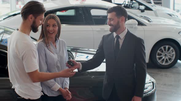 Successful Deal Between Car Salesman and Young Couple in Car Dealership. alt