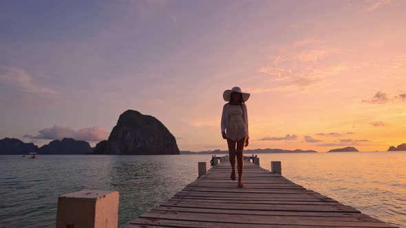 Woman Walking Along Wooden Pier As Sun Sets Over The Ocean alt